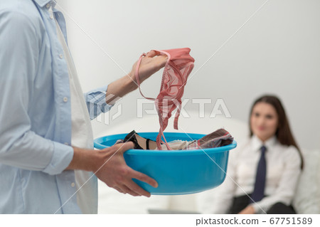 Man holding up his wifes lingerie. Selective focus on male hand picking out light pink bra out of laundry basin. Householder husband concept. Close up shot. Woman on bed blurred in background Man holding up his wifes lingerie. Selective focus on male hand picking out light pink bra out of laundry basin. Householder husband concept. Close up shot. Woman on bed blurred in background 67751589