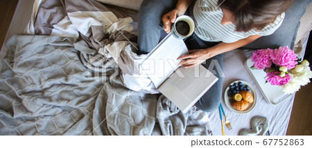 Woman sitting in bed reading a book and having breakfast. Top view 67752863
