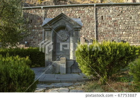 Monument is a source of water, carved from basalt, in a park near a stone wall located around Geghard Monastery in Armenia Monument is a source of water, carved from basalt, in a park near a stone wall located around Geghard Monastery in Armenia 67753887