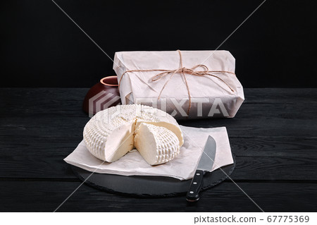 Adyghe cheese cut off a triangular piece. Nearby are three cheese heads packed in paper. On a wooden board. Background gray concrete Adyghe cheese cut off a triangular piece. Nearby are three cheese heads packed in paper. On a wooden board. Background gray concrete 67775369