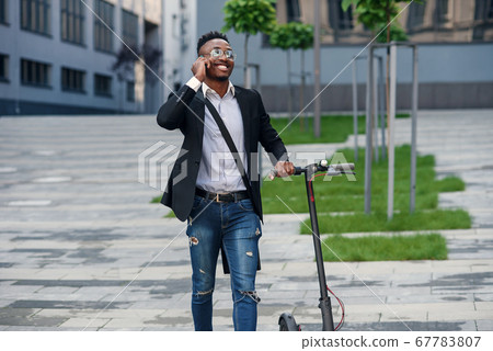 Smiling businessman with electric scooter standing near modern business building talking on phone. Smiling businessman with electric scooter standing near modern business building talking on phone. 67783807
