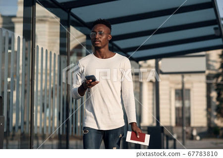 Young black male tourist with suitcase and passport in hands on bus of subway stop using internet maps in smartphone. Tourist finding a right way on smartphone in new city. Young black male tourist with suitcase and passport in hands on bus of subway stop using internet maps in smartphone. Tourist finding a right way on smartphone in new city. 67783810