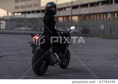 Adorable girl in black tight body suit and full-face helmet rides on stylish motorcycle at urban outdoors parking in evening. Freedom and active lifestyle concept. Totally black. 67784349