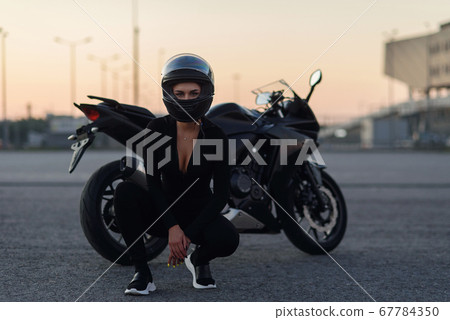 Biker woman in black leather jacket and full face helmet sits near stylish sports motorcycle at urban parking. Traveling and active lifestyle concept. 67784350
