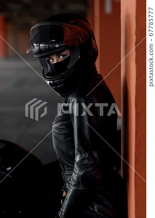 Stylish young woman motorcycle rider with beautiful eyes in black protective gear and full-face helmet near her bike on underground parking. Extremal and freedom concept. Stylish young woman motorcycle rider with beautiful eyes in black protective gear and full-face helmet near her bike on underground parking. Extremal and freedom concept. 67785777