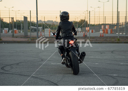 Back view of stylish woman in black leather jacket, pants and protective helmet rides on sports motorcycle at urban outdoors parking. Back view of stylish woman in black leather jacket, pants and protective helmet rides on sports motorcycle at urban outdoors parking. 67785819