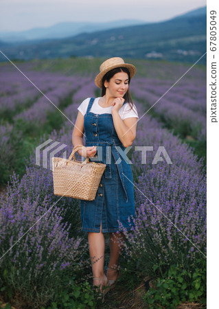 Attractive young woman walking in summer lavender field. Model dressed in denim sundress with straw hat and bag Attractive young woman walking in summer lavender field. Model dressed in denim sundress with straw hat and bag 67805049