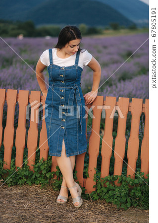 Attractive young woman walking in summer lavender field. Model dressed in denim sundress 67805191