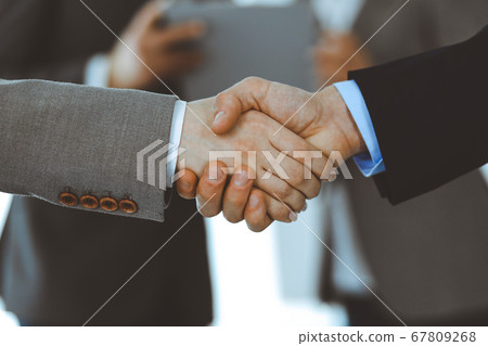 Business people shaking hands while standing with colleagues after meeting or negotiation, close-up. Group of unknown businessmen and women in modern office. Teamwork, partnership and handshake 67809268