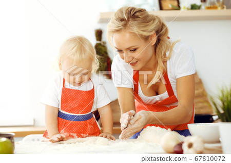 Little girl and her blonde mom in red aprons playing and laughing while kneading the dough in kitchen. Homemade pastry for bread, pizza or bake cookies. Family fun and cooking concept 67810072