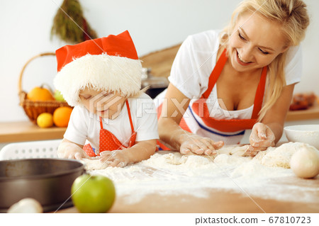 Little girl and her blonde mom in red aprons playing and laughing while kneading the dough in kitchen. Homemade pastry for bread, pizza or bake cookies. Family fun and cooking concept 67810723
