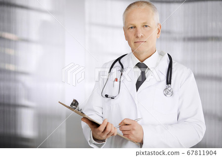 Elderly confident doctor standing straight and using clipboard in clinic. Perfect medical service. Medicine and healthcare concept 67811904