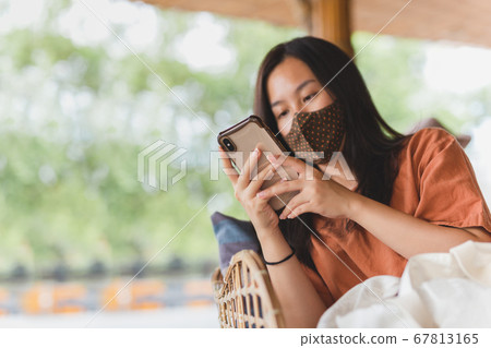 Woman in face mask using her mobile phone while sitting in a chair. Woman in face mask using her mobile phone while sitting in a chair. 67813165