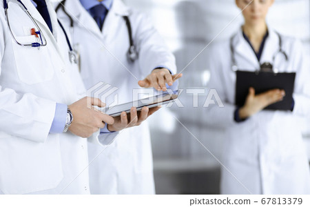 Group of unknown doctors use a computer tablet to check up some medical names records, while standing in a hospital office. Physicians ready to examine and help patients. Medical help, insurance in 67813379