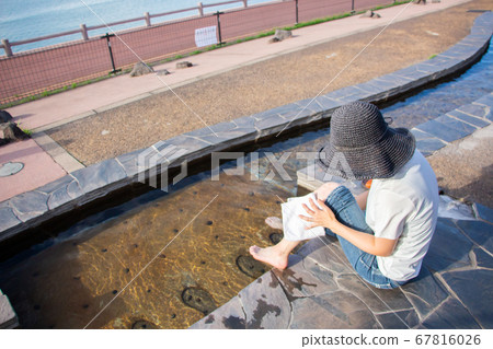在鹿兒島縣塔魯米市的路邊車站Yusurikan，一名身穿白襯衣，戴著黑帽子的婦女用洗腳盆擦腳。 67816026