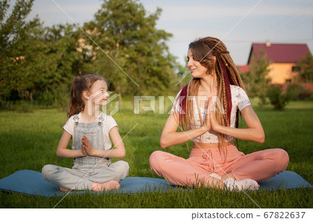 Young mother with dreadlocks and little daughter are doing yoga exercises in lotus position on grass in the park at the day time. Concept of friendly family and of summer vacation. 67822637