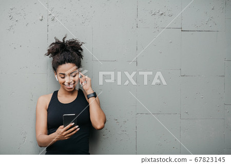 Sport gadgets. African american girl with fitness tracker and headphones looks at smartphone and listens to music Sport gadgets. African american girl with fitness tracker and headphones looks at smartphone and listens to music 67823145