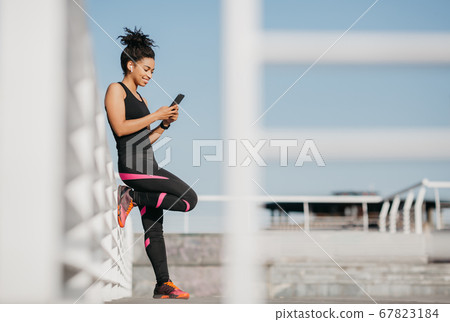 African american girl in sport uniform leans on wall with wireless headphones and looks at phone African american girl in sport uniform leans on wall with wireless headphones and looks at phone 67823184