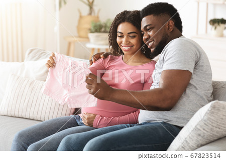 Smiling black expecting couple holding baby clothes Smiling black expecting couple holding baby clothes 67823514