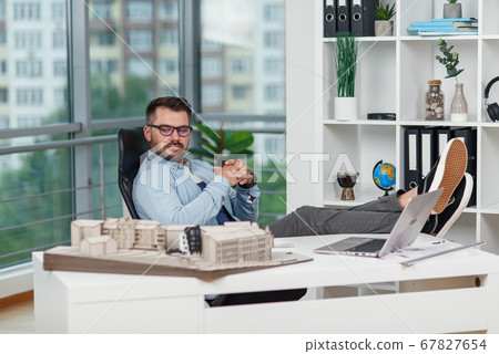 Handsome experienced office worker in casual clothes laid his feet on workspace table while dreaming about rest or vacations. Joyful architect relax on the workplace successfully completing his Handsome experienced office worker in casual clothes laid his feet on workspace table while dreaming about rest or vacations. Joyful architect relax on the workplace successfully completing his 67827654