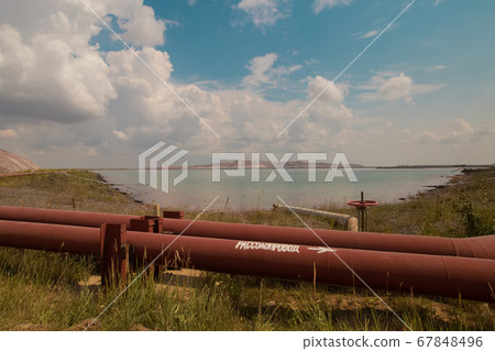 Soligork salt mine slagheap mountains and scenic nature with green grass and dramatic cloudy sky in summer day. A technical water lake, industrial metal constructions, brine pipeline, old rusty pipes 67848496