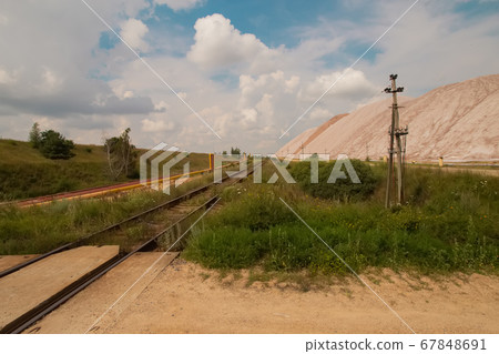 Soligork salt mine slagheap mountains and scenic beautiful nature with green grass and dramatic cloudy sky in hot summer day. Old rusty railway path and industrial metal constructions, pipes. Soligork salt mine slagheap mountains and scenic beautiful nature with green grass and dramatic cloudy sky in hot summer day. Old rusty railway path and industrial metal constructions, pipes. 67848691