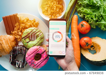 Calories counting and food control concept. woman using Calorie counter application on her smartphone with fresh vegetables, dessert and donuts on dining table Calories counting and food control concept. woman using Calorie counter application on her smartphone with fresh vegetables, dessert and donuts on dining table 67849838