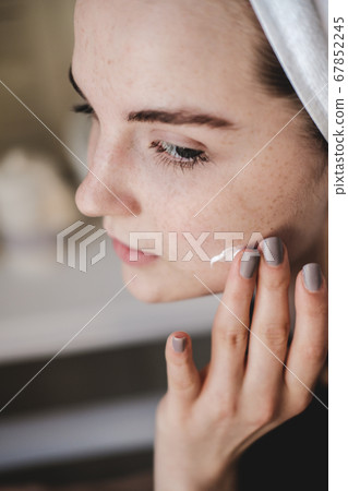 Young beautiful woman with towel on her head after shower in the bathroom applies moisturizer to her face. The concept of female morning skin and hair care procedures. Young beautiful woman with towel on her head after shower in the bathroom applies moisturizer to her face. The concept of female morning skin and hair care procedures. 67852245