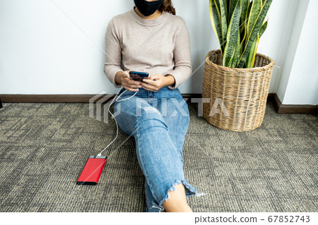 young woman sitting against a wall holding a smartphone while charging a smartphone battery 67852743