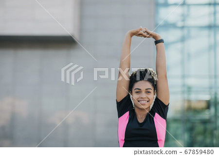 Smiling african american girl in sportswear with wireless headphones and fitness tracker doing stretching for hands Smiling african american girl in sportswear with wireless headphones and fitness tracker doing stretching for hands 67859480