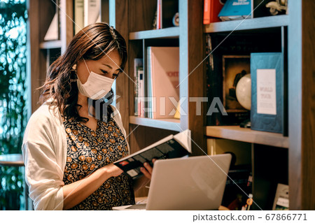 Young woman in mask working desk in front of bookshelf in office Young woman in mask working desk in front of bookshelf in office 67866771