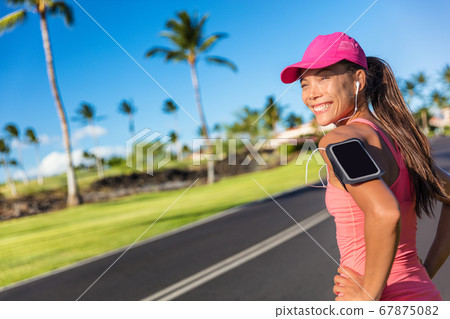 Fitness motivation running music woman runner. Happy jogging girl listening to smartphone app with earphones. Asian athlete wearing cap and armband smart phone holder outdoor on city road 67875082