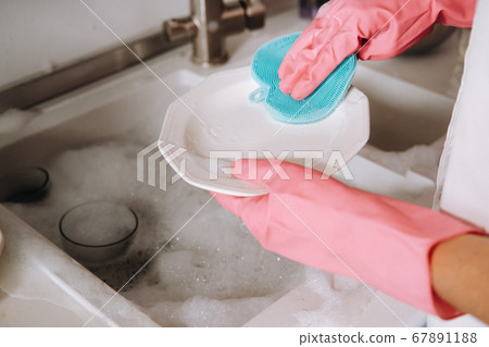 housewife girl in pink gloves washes dishes by hand in the sink with detergent. The girl cleans the house and washes dishes in gloves at home. housewife girl in pink gloves washes dishes by hand in the sink with detergent. The girl cleans the house and washes dishes in gloves at home. 67891188