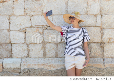 Beautiful young female tourist woman wearing big straw hat, taking self portrait selfie, standing in front of old textured stone wall at old Mediterranean town Beautiful young female tourist woman wearing big straw hat, taking self portrait selfie, standing in front of old textured stone wall at old Mediterranean town 67903193