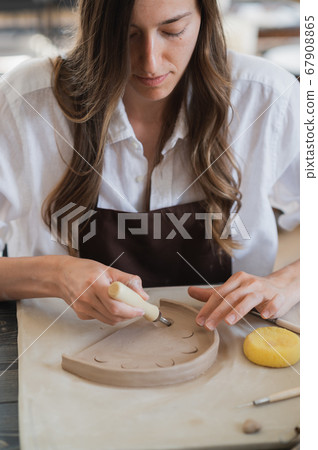 Craftsman hands connect the clay parts made of raw clay, master fingers work with pieces of clay, female sits at a workshop behind the table 67908865
