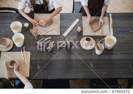 Top view of female hands sculpting different clay products. Big wooden table with pottery tools on a workshop. 67908870