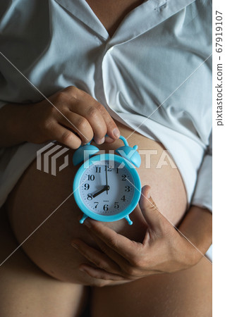 High angle of crop pregnant female holding alarm clock on belly while waiting for baby. 67919107