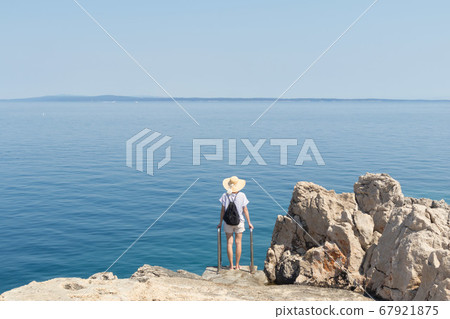 Woman traveler wearing straw summer hat and backpack, standing at edge of the rocky cliff looking at big blue sea and islands in on the horizon Woman traveler wearing straw summer hat and backpack, standing at edge of the rocky cliff looking at big blue sea and islands in on the horizon 67921875