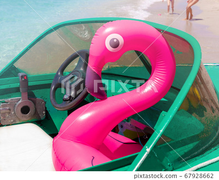 Inflatable pink flamingo sitting inside the green powerboat on the white seat with legs running on the sand beach on the background on sunny summer day by the sea. Family vacation, holidays, fun, joy Inflatable pink flamingo sitting inside the green powerboat on the white seat with legs running on the sand beach on the background on sunny summer day by the sea. Family vacation, holidays, fun, joy 67928662
