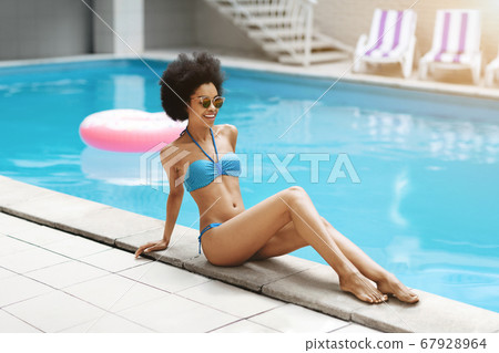 Attractive black lady in bikini sitting near swimming pool at tropical summer resort 67928964