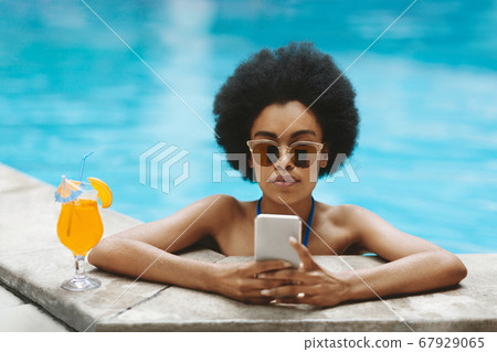 Gorgeous black lady with tropical cocktail using smartphone at outdoor pool 67929065