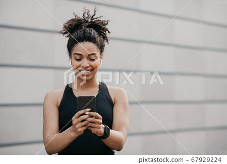 Great mood, music and sport. Happy african american athletic girl in sportswear with fitness tracker uses smartphone 67929224