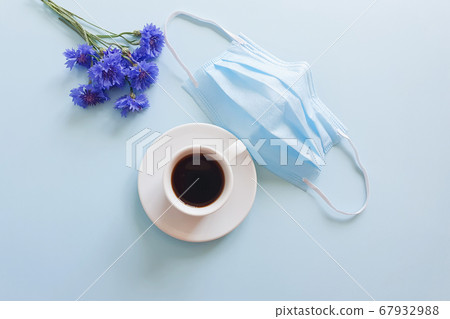 Medical protective face mask from coronavirus Covid-19, cup of coffee and blue flowers on a pastel blue background. Biological concept of life in a new reality. Top view, flat lay lifestyle. 67932988