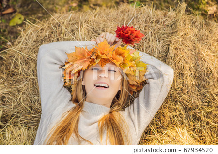 Outdoors lifestyle close up portrait of charming blonde young woman wearing a wreath of autumn leaves on a haystack. Wearing stylish knitted pullover. Wreath of maple leaves Outdoors lifestyle close up portrait of charming blonde young woman wearing a wreath of autumn leaves on a haystack. Wearing stylish knitted pullover. Wreath of maple leaves 67934520