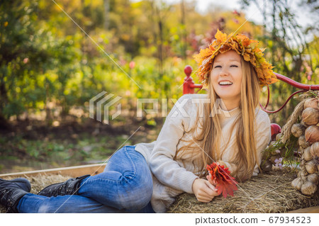 Outdoors lifestyle close up portrait of charming blonde young woman wearing a wreath of autumn leaves on a haystack. Wearing stylish knitted pullover. Wreath of maple leaves Outdoors lifestyle close up portrait of charming blonde young woman wearing a wreath of autumn leaves on a haystack. Wearing stylish knitted pullover. Wreath of maple leaves 67934523
