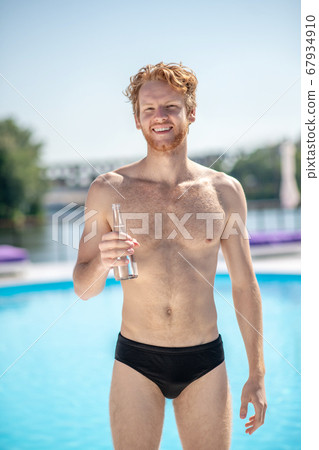 Muscular man with bottle of water standing near pool 67934910