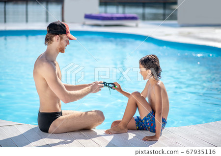 Man and boy sitting opposite each other near swimming pool Man and boy sitting opposite each other near swimming pool 67935116