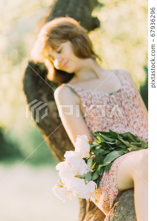 Beautiful young Caucasian blond woman in light dress holding bouquet of white peonies, walking in summer field or garden in sunset. Woman with flowers outdoors 67936296