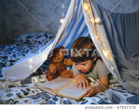 Little boy reads book. Toddler plays in tent made of linen sheet on bed. Cozy evening with favorite book. Little boy reads book. Toddler plays in tent made of linen sheet on bed. Cozy evening with favorite book. 67937378