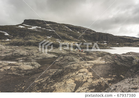 Tourists with backpacks walk across a rocky plain to the famous landmark - Troll tongue rock or Trolltunga. Cold and windy summer in Norway. Tourists with backpacks walk across a rocky plain to the famous landmark - Troll tongue rock or Trolltunga. Cold and windy summer in Norway. 67937380